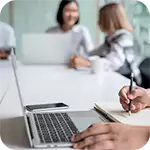 Business professionals collaborating in a meeting, one taking notes on a laptop and notebook for a Regulatory Requirement review.
